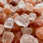 A close-up of coarse Himalayan pink salt crystals on a dark wooden table, showcasing their natural pink color.